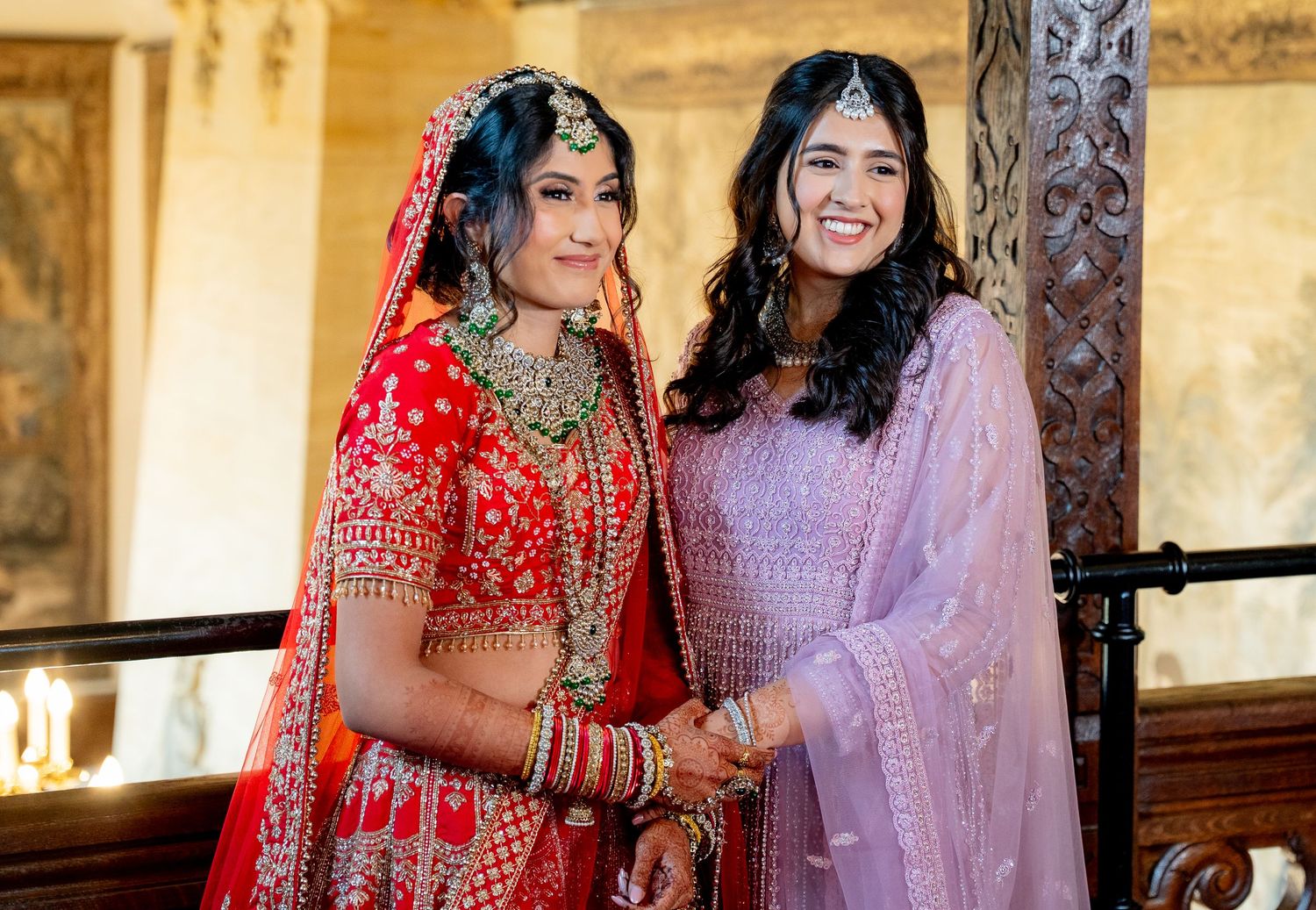 Two women in traditional Indian attire pose together, one in a red bridal lehenga with gold jewelry, the other in a lavender suit.