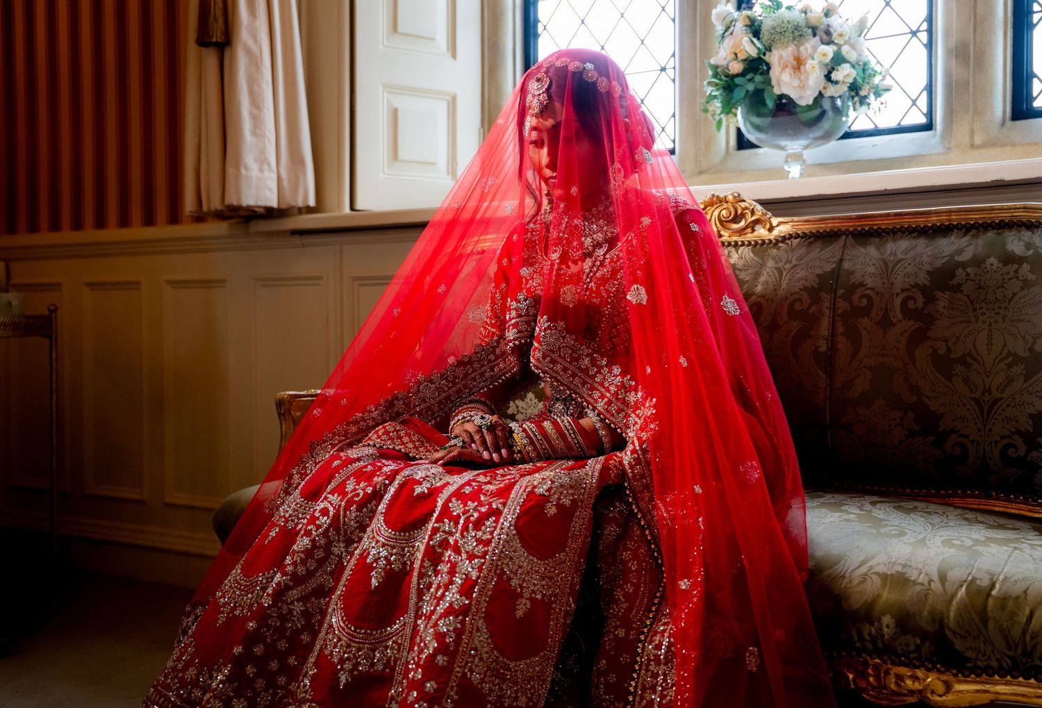 Bride in stunning red bridal lehenga with veil sits by window in ornate wedding attire.