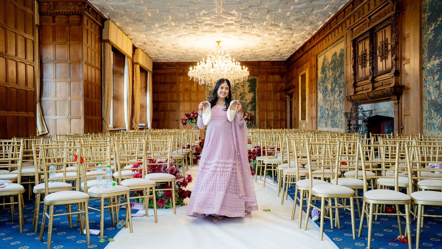 Woman in pink lehenga standing in elegant wedding venue with gold chairs.