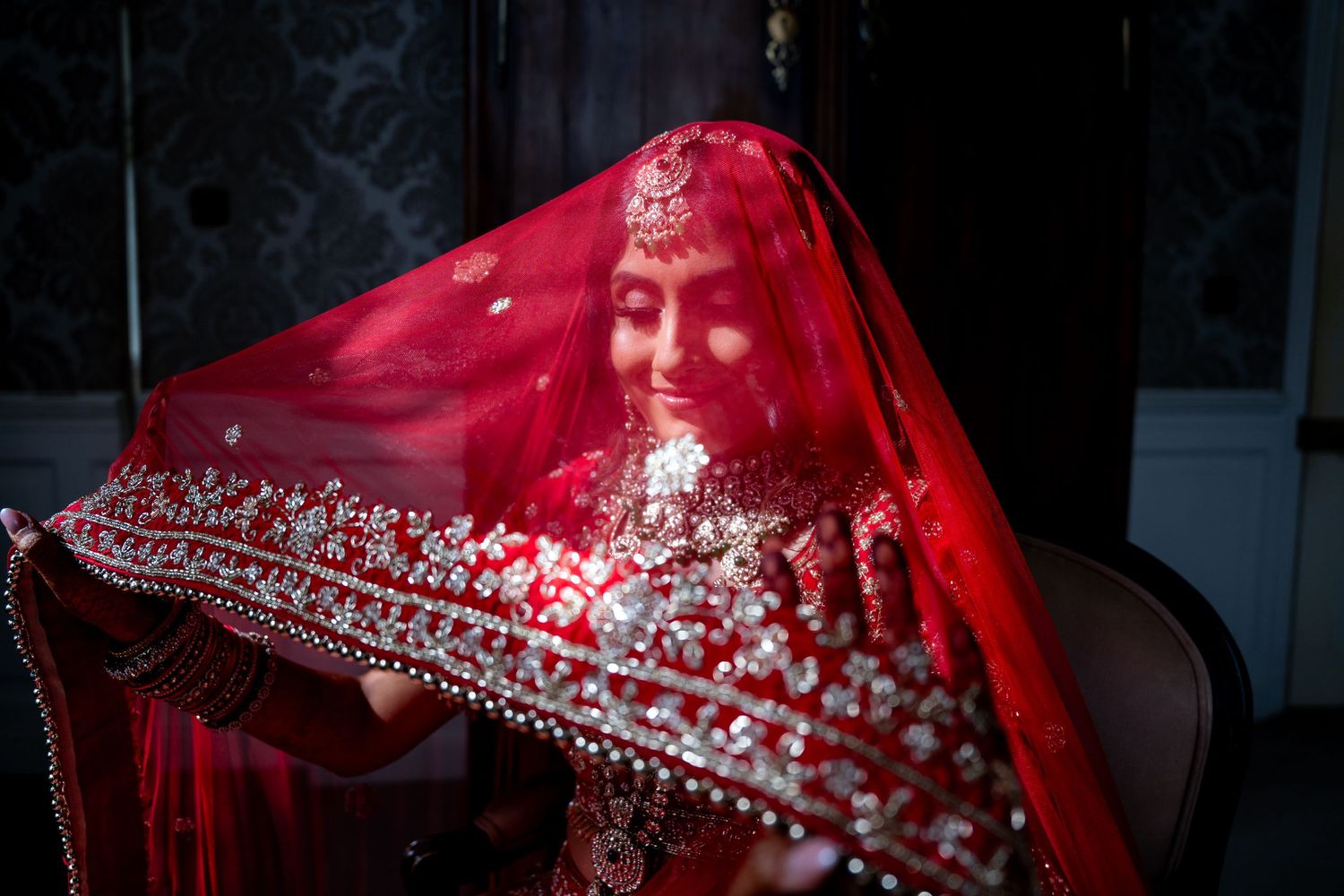 Indian bride in red embroidered veil smiling softly during traditional wedding preparation.
