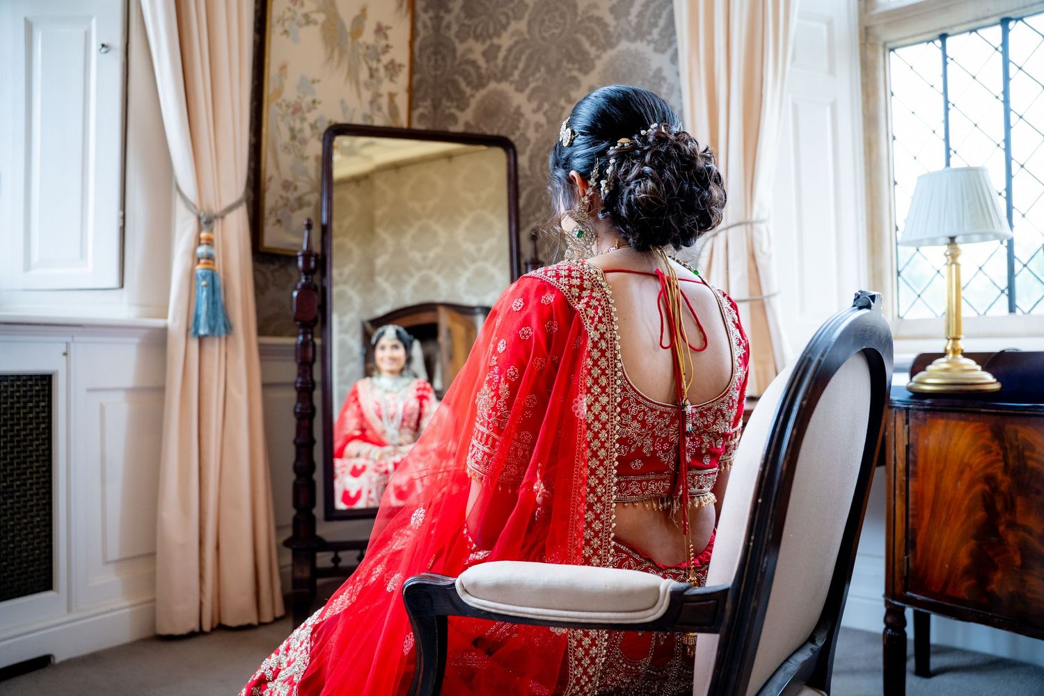 Indian bride in red embroidered lehenga seated before mirror reflecting her elegant bridal look.