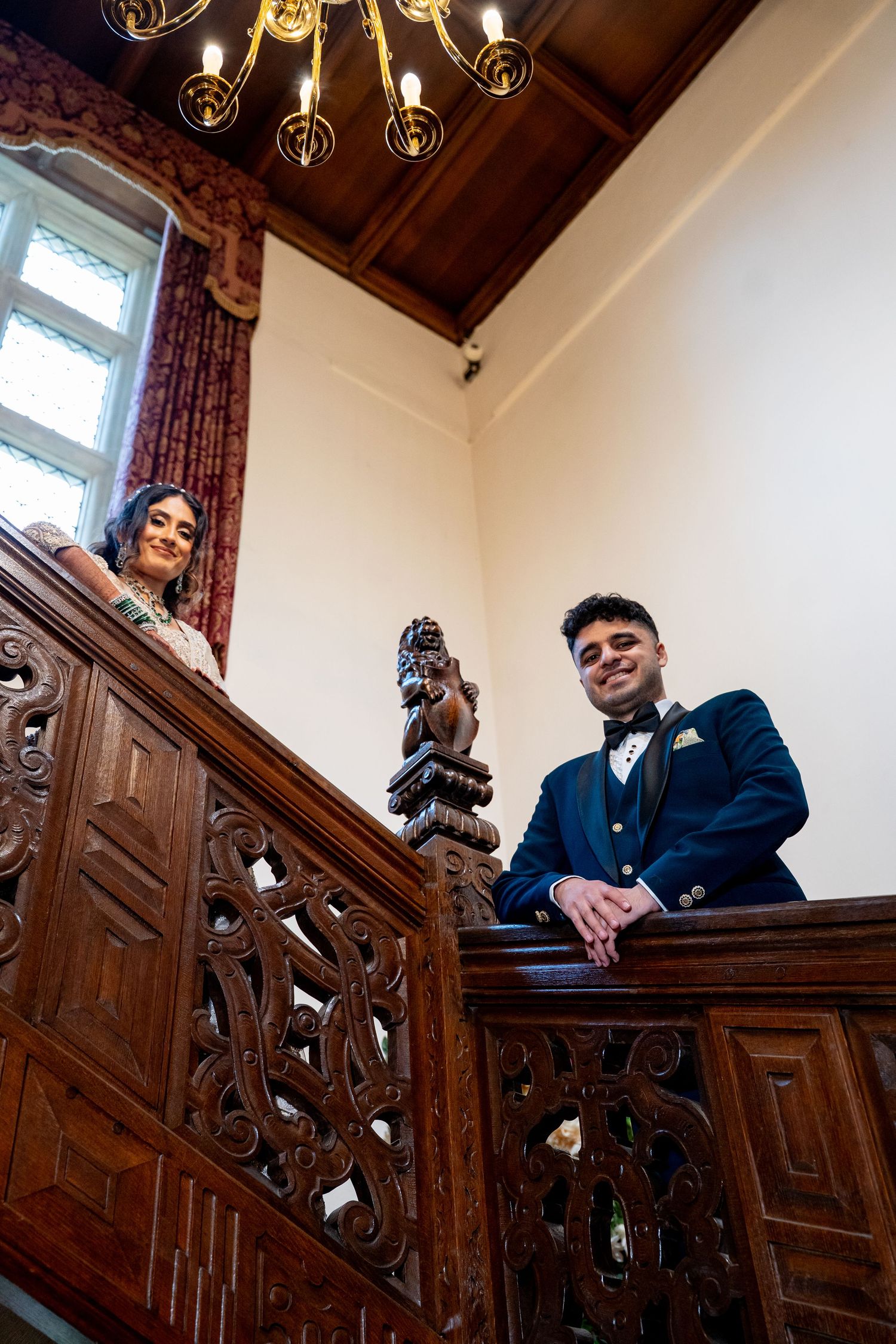 Bride and groom lean over ornate carved wooden staircase railing in a grand historic venue.