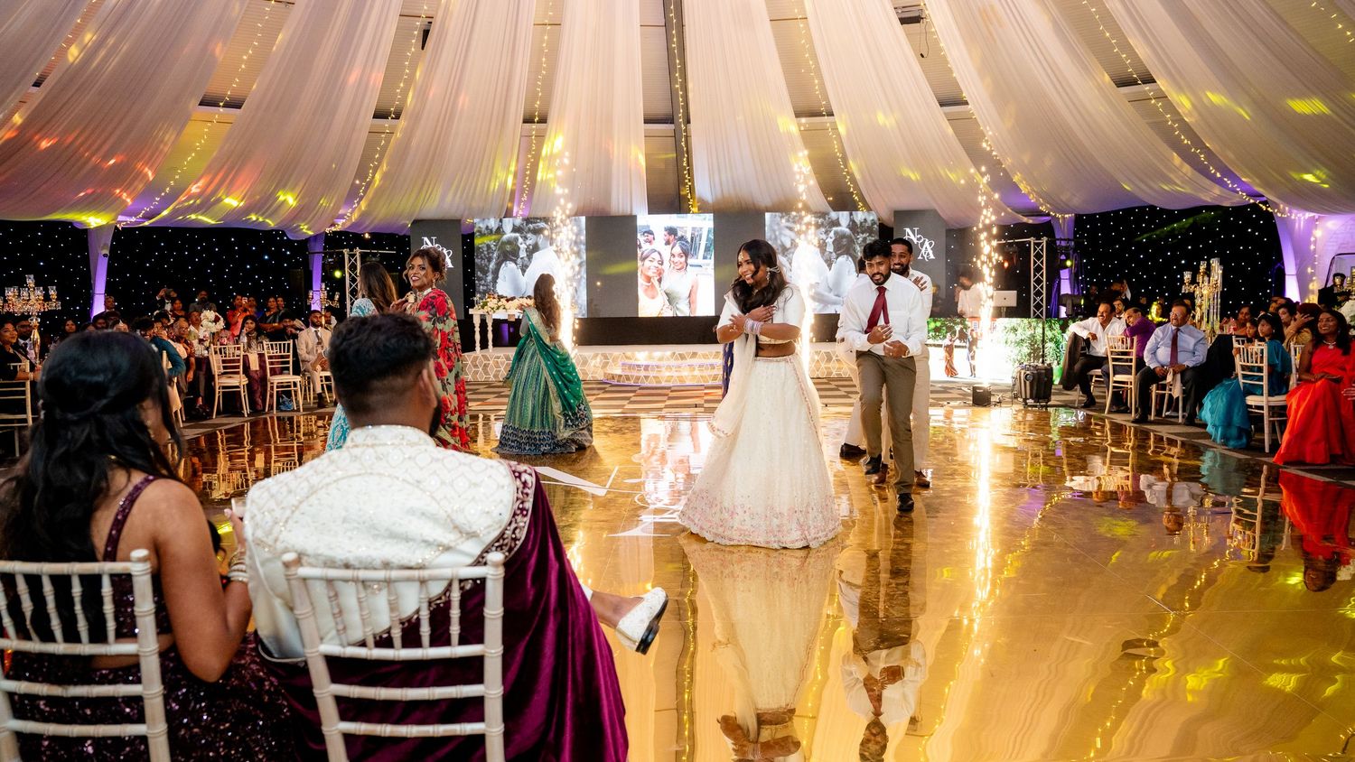 Elegant wedding reception under a draped tent with couple dancing on a gold reflective floor surrounded by guests.
