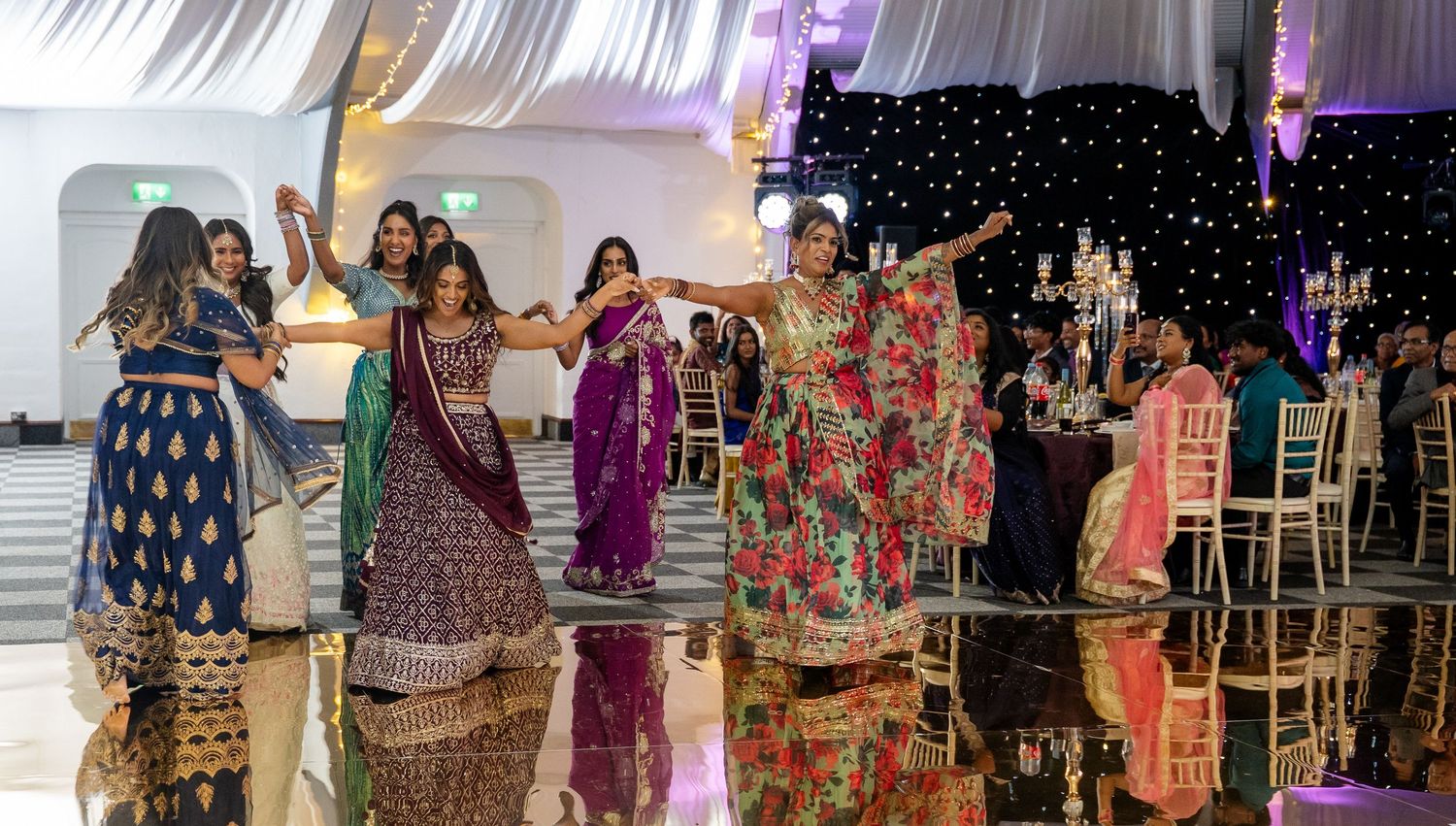 Group of women in vibrant Indian lehengas and sarees dancing together at a South Asian wedding reception.