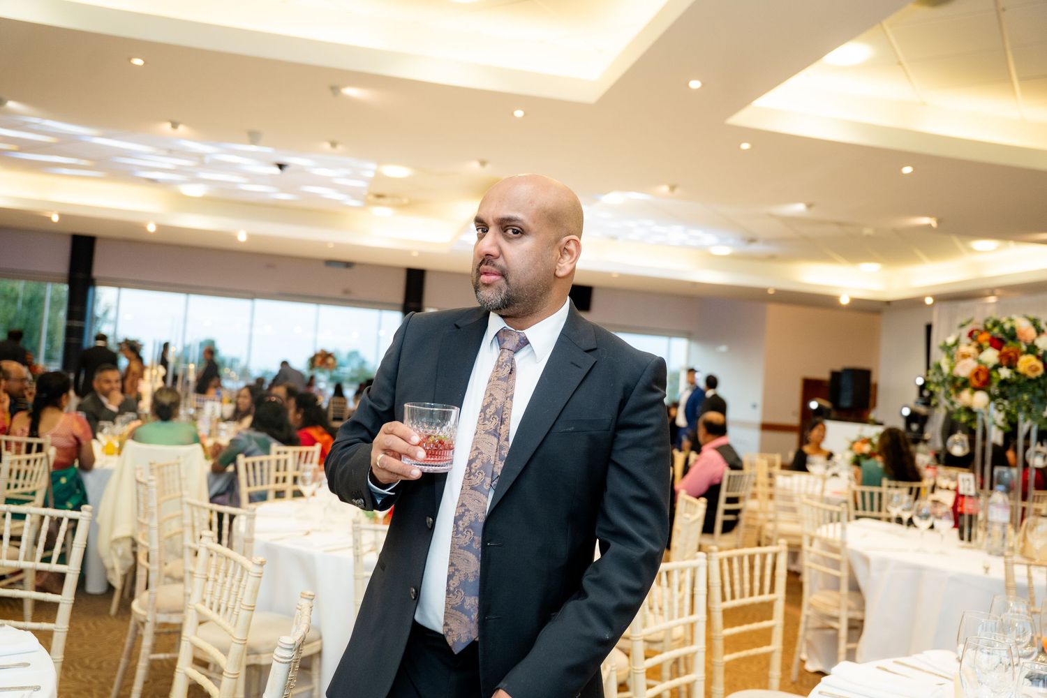 Man in dark suit holds a drink while standing in a bright decorated wedding banquet hall.
