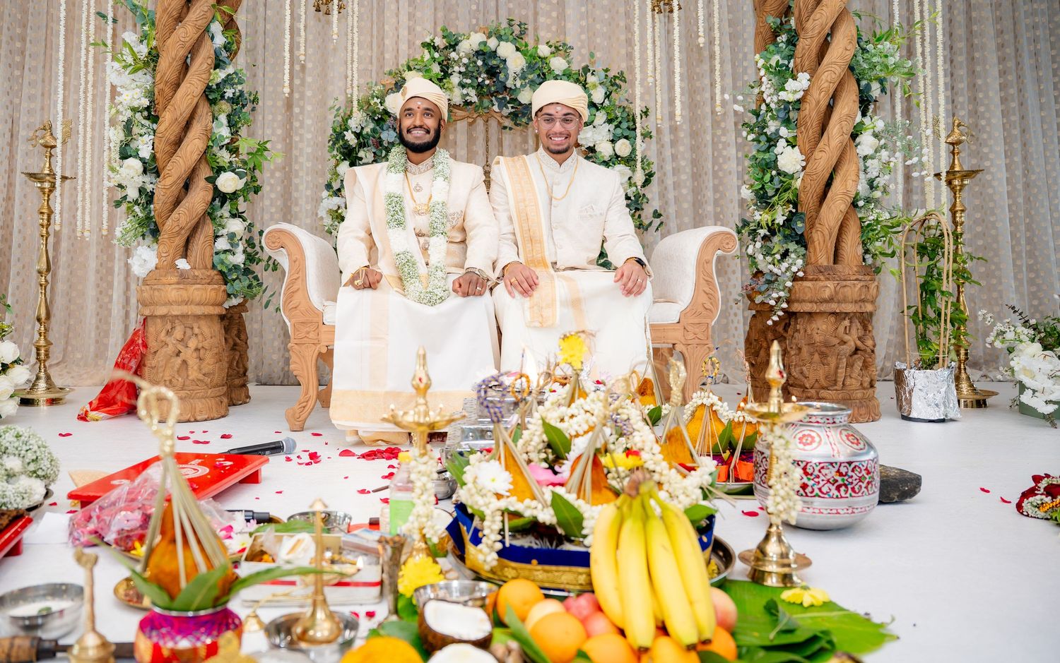 Two grooms in white sherwanis sit on decorated mandap stage at traditional South Indian wedding ceremony.