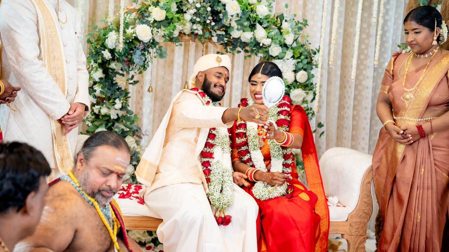 Groom holds mirror for bride during traditional South Indian Hindu wedding ceremony with floral backdrop.