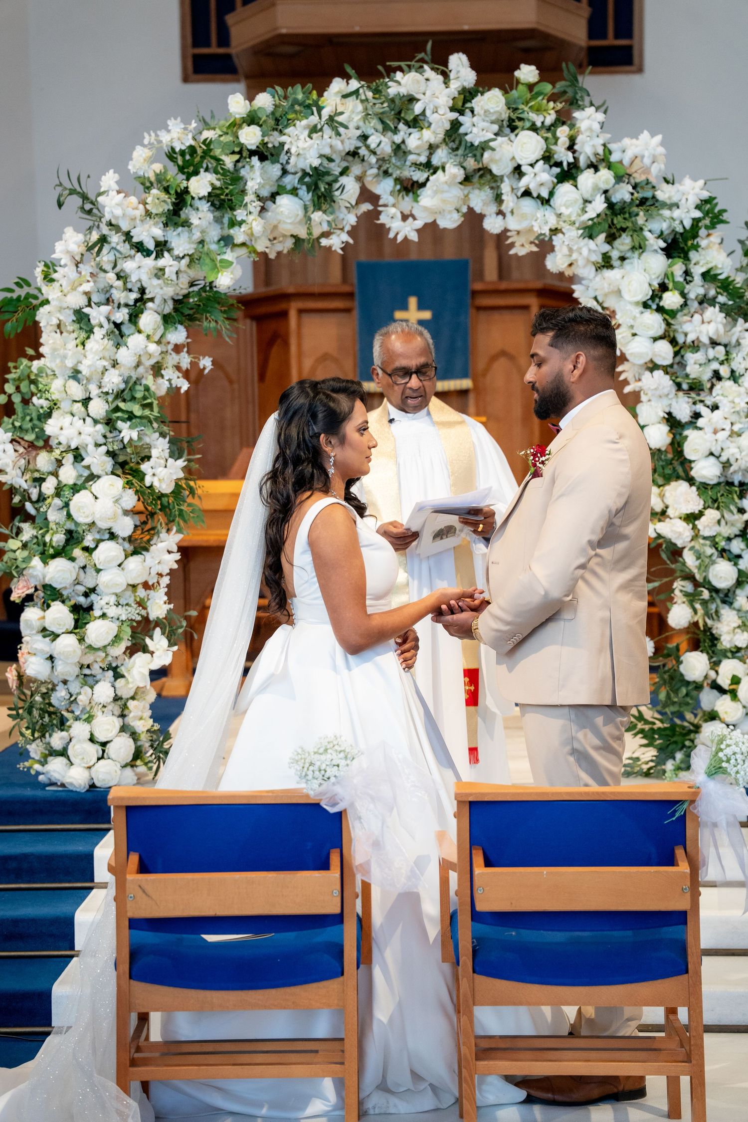 Bride and groom hold hands exchanging vows under white floral arch with officiant behind them.
