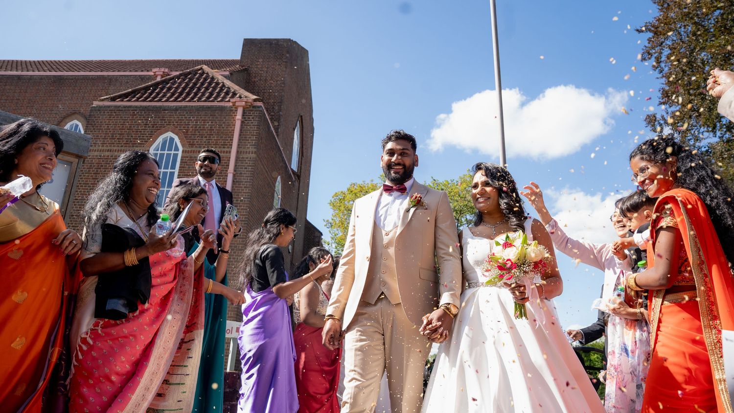 Indian wedding couple exits church as guests toss flower petals in joyful celebration outdoors.