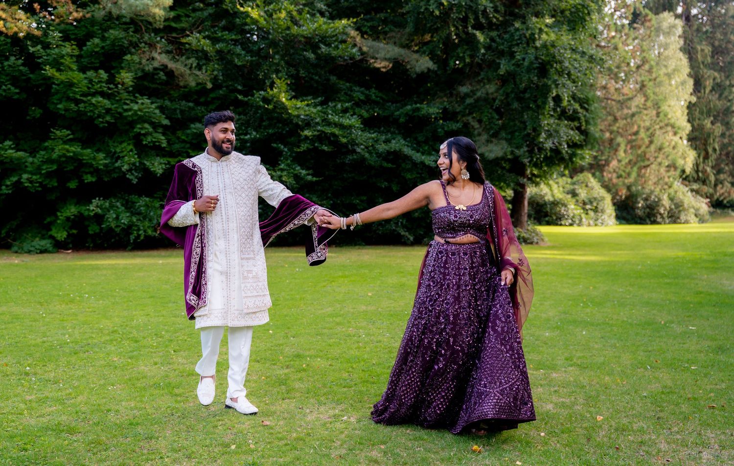 Indian wedding couple holding hands walking on green lawn, bride in purple lehenga, groom in white sherwani.