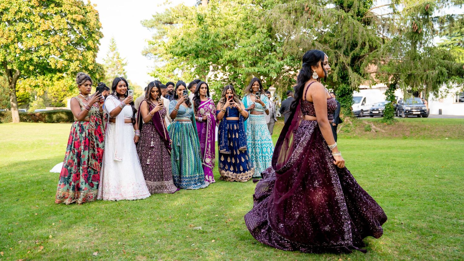 A woman in a stunning purple lehenga twirls on green lawn while guests in colorful Indian attire clap and photograph her.