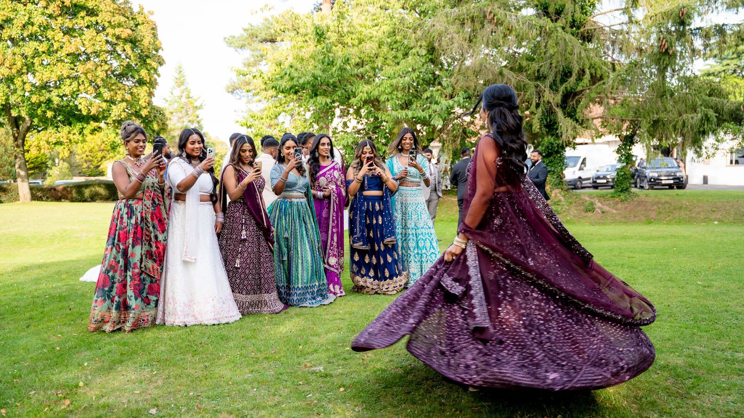 A bride in a purple lehenga twirls while guests in colorful Indian attire cheer on a lawn.