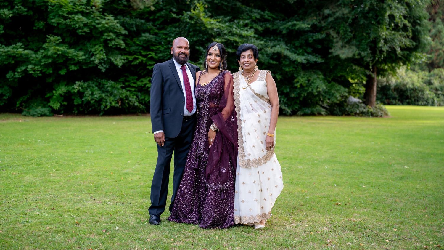Family portrait of three people in traditional South Asian wedding attire on a green lawn.