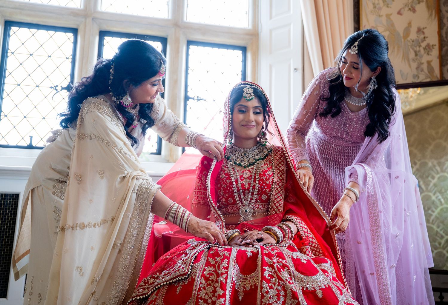 Two women helping Indian bride in red lehenga with jewelry and finishing touches before wedding.