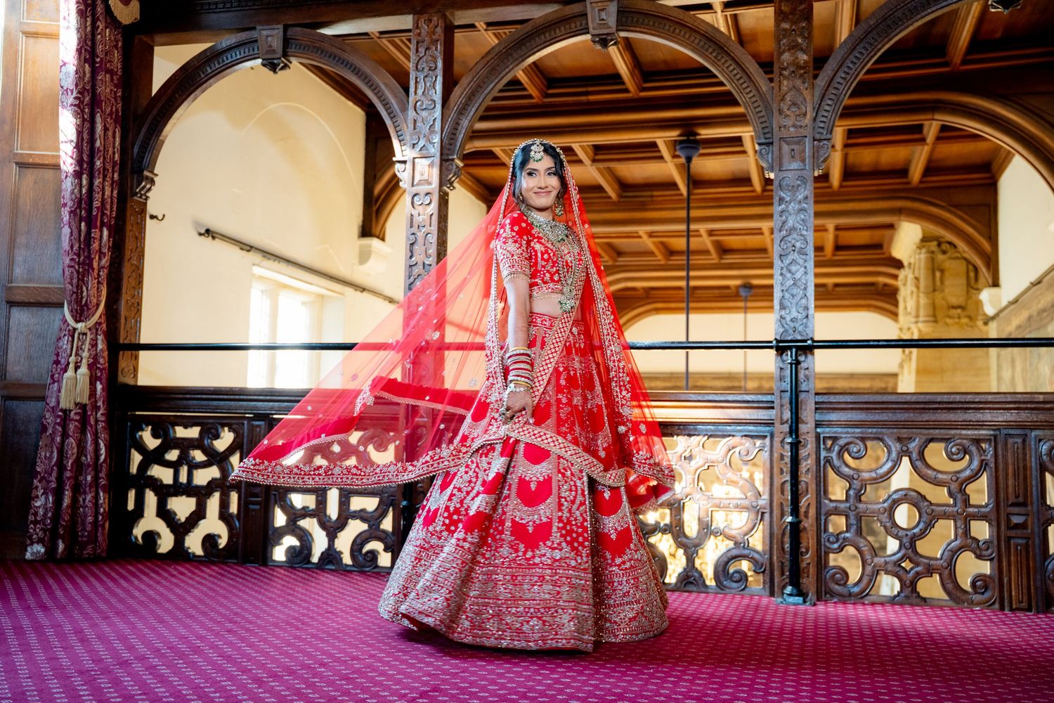 A bride in a stunning red embroidered lehenga with dupatta poses in an ornate wooden hall.