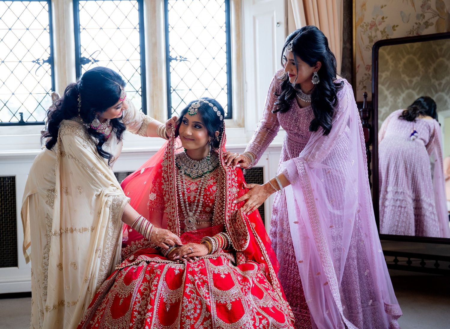Two women help a bride in a red lehenga with jewelry during Indian wedding preparations.