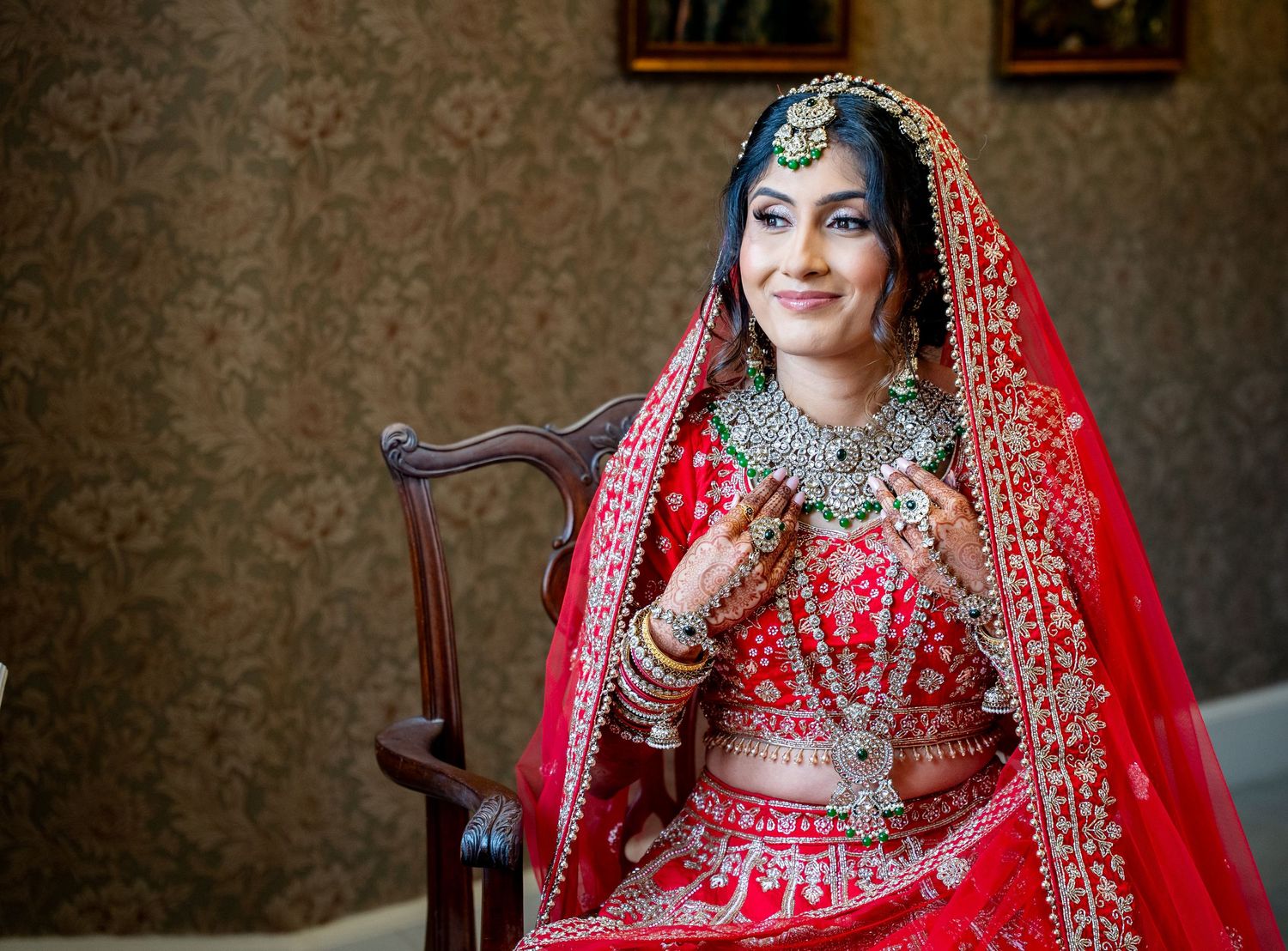 Indian bride in red lehenga with gold jewelry and dupatta seated on chair for traditional wedding portrait.