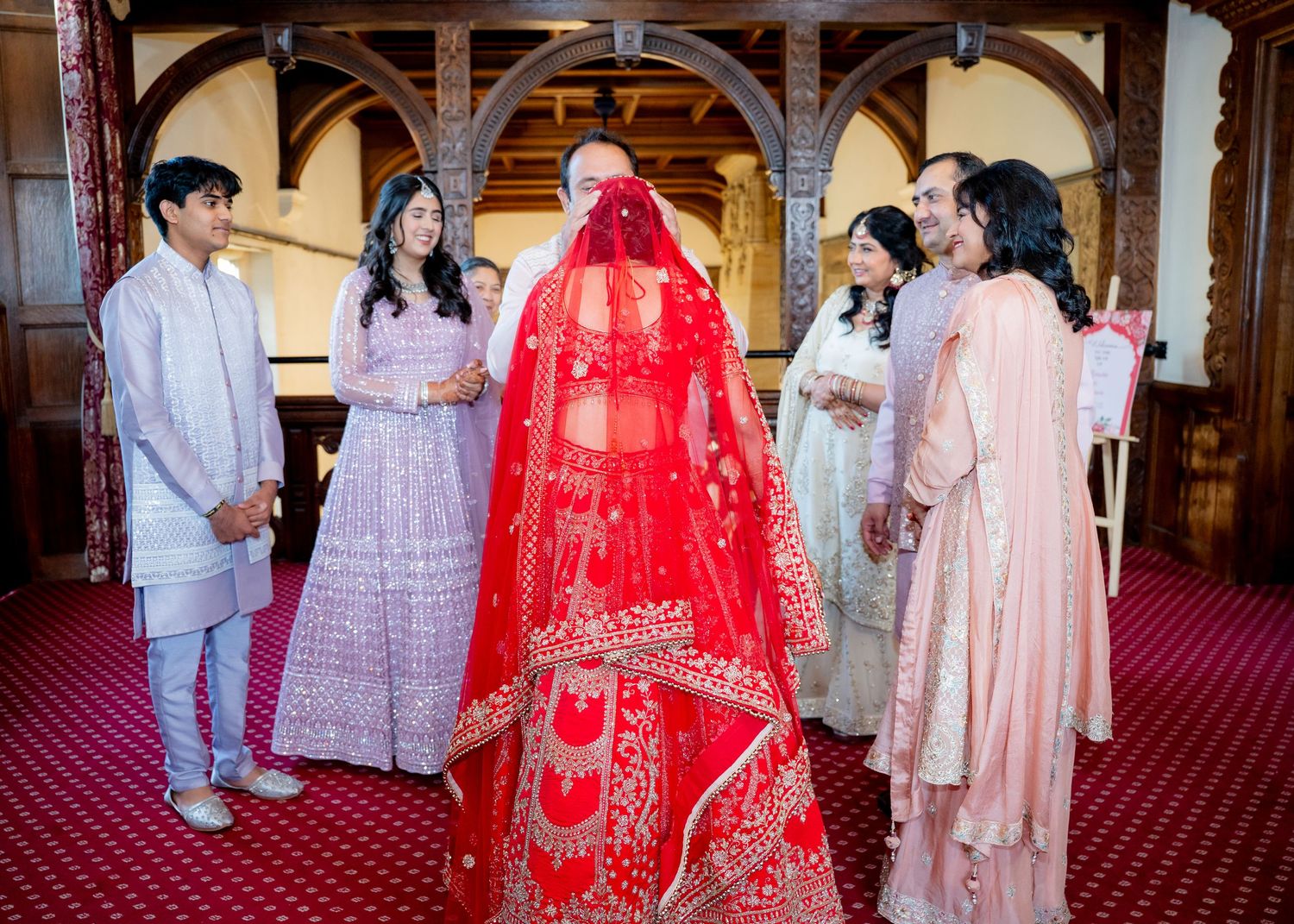 Indian bride in red lehenga sharing a joyful moment with family members in grand hall.