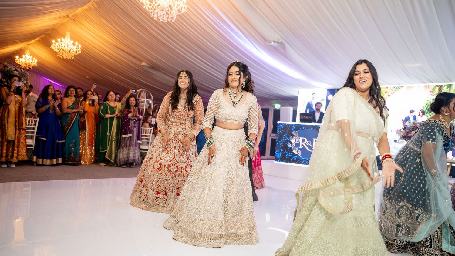 Women in vibrant traditional Indian lehengas dancing at an elegant wedding reception in a decorated banquet hall.