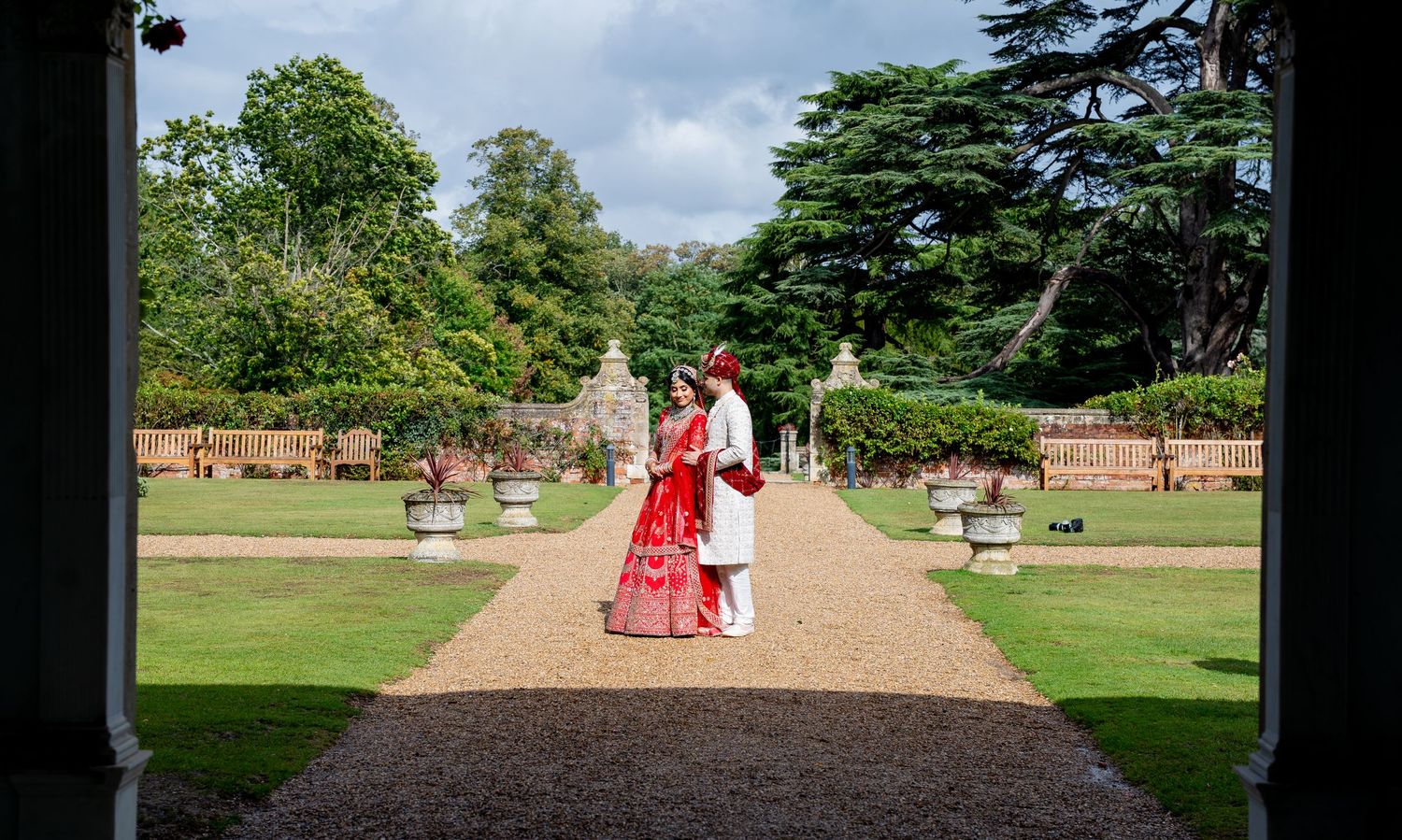 Indian bride and groom in red and white attire stand facing each other in a formal English garden.