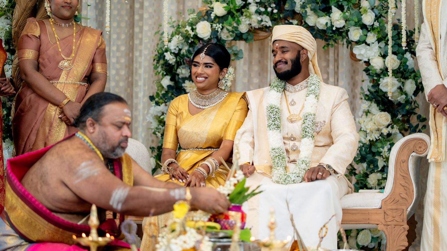 Hindu wedding ceremony with bride in gold saree and groom in white sherwani seated at mandap with priest.