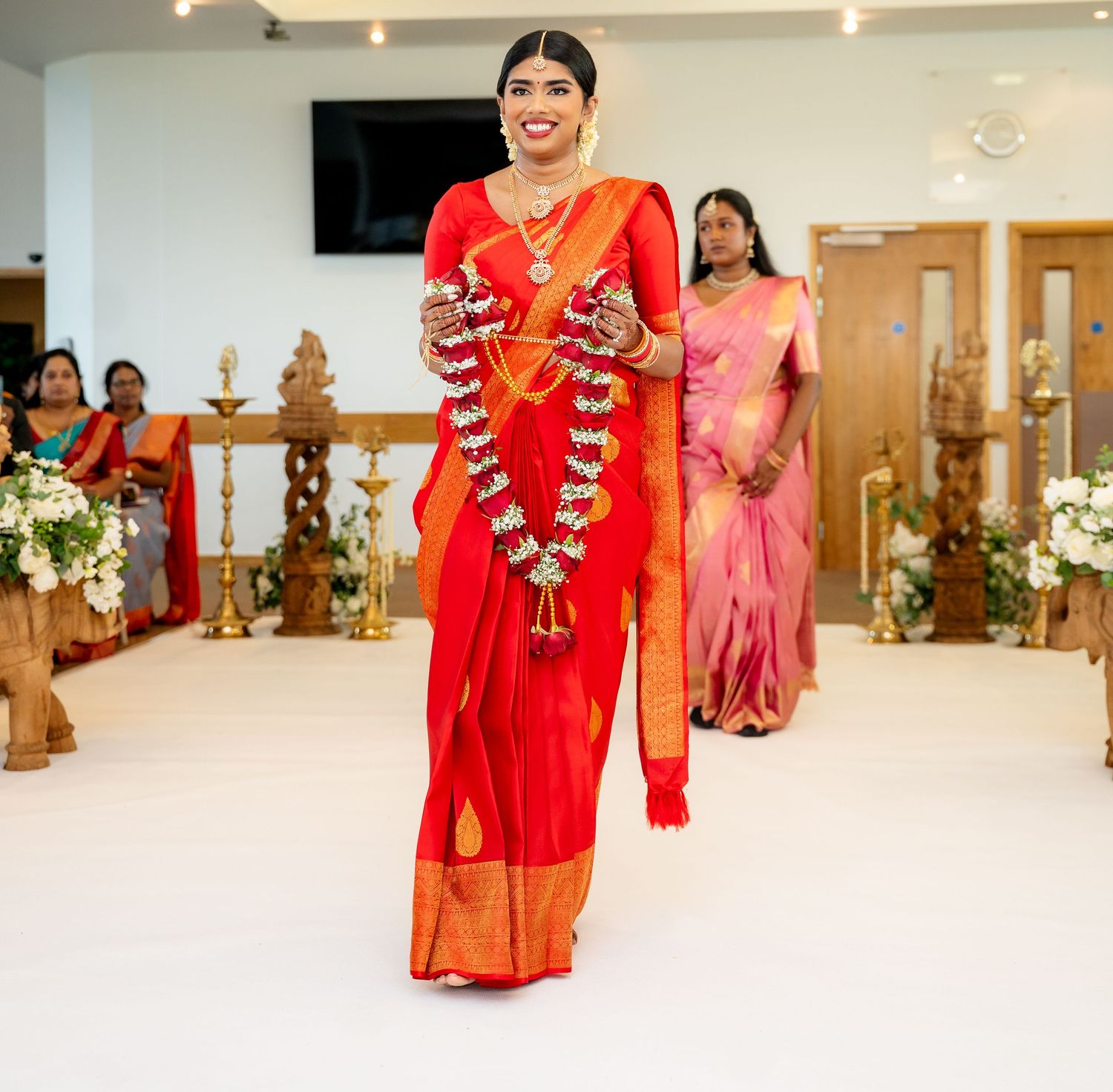 Smiling bride in red Kanjivaram saree holds jasmine garlands while walking down wedding aisle.