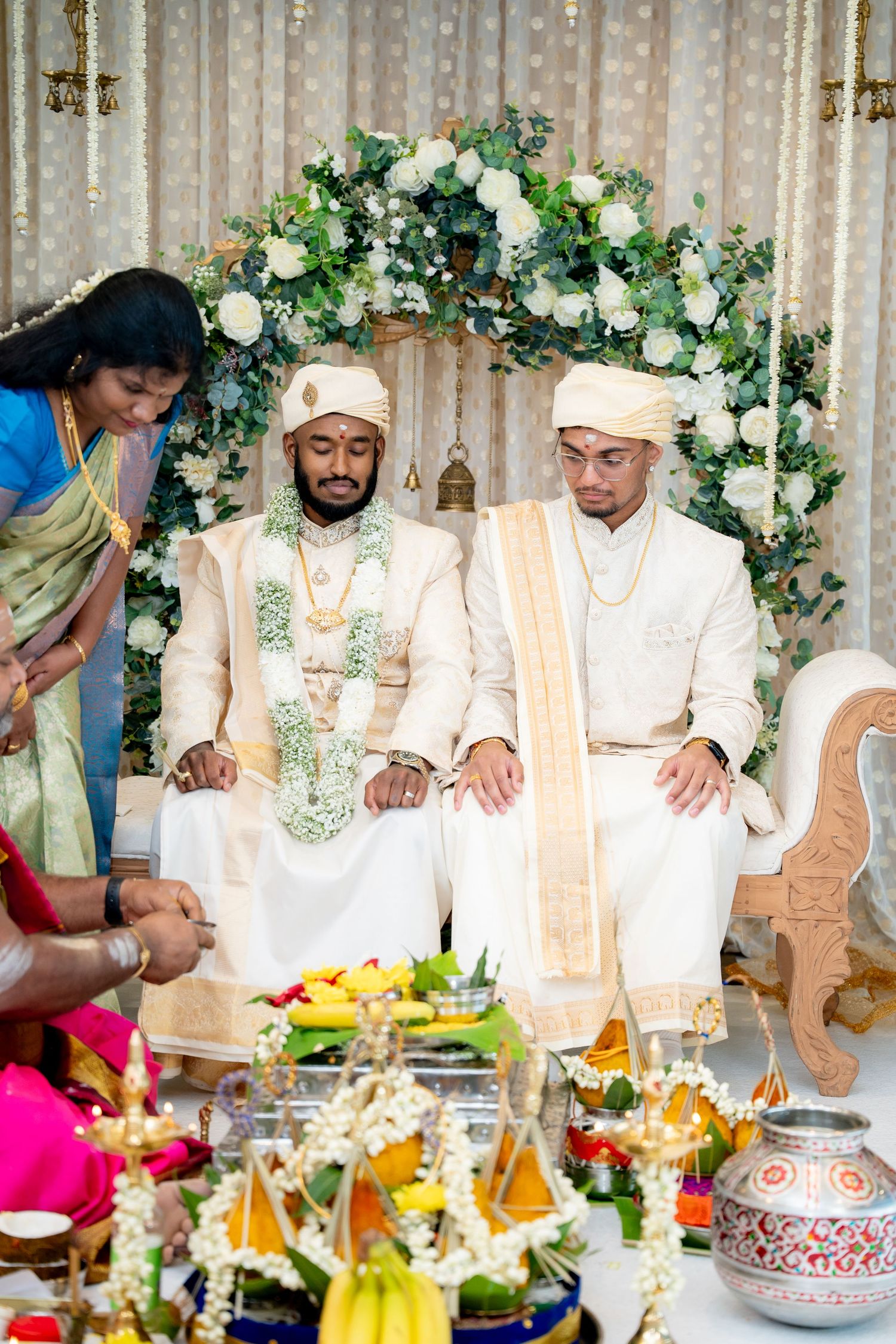 Two grooms in white sherwanis and turbans sit at a traditional South Asian wedding ceremony with floral decorations.