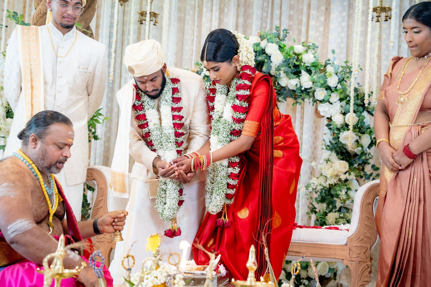 Bride in red saree and groom perform traditional Hindu wedding rituals with priest at mandap.