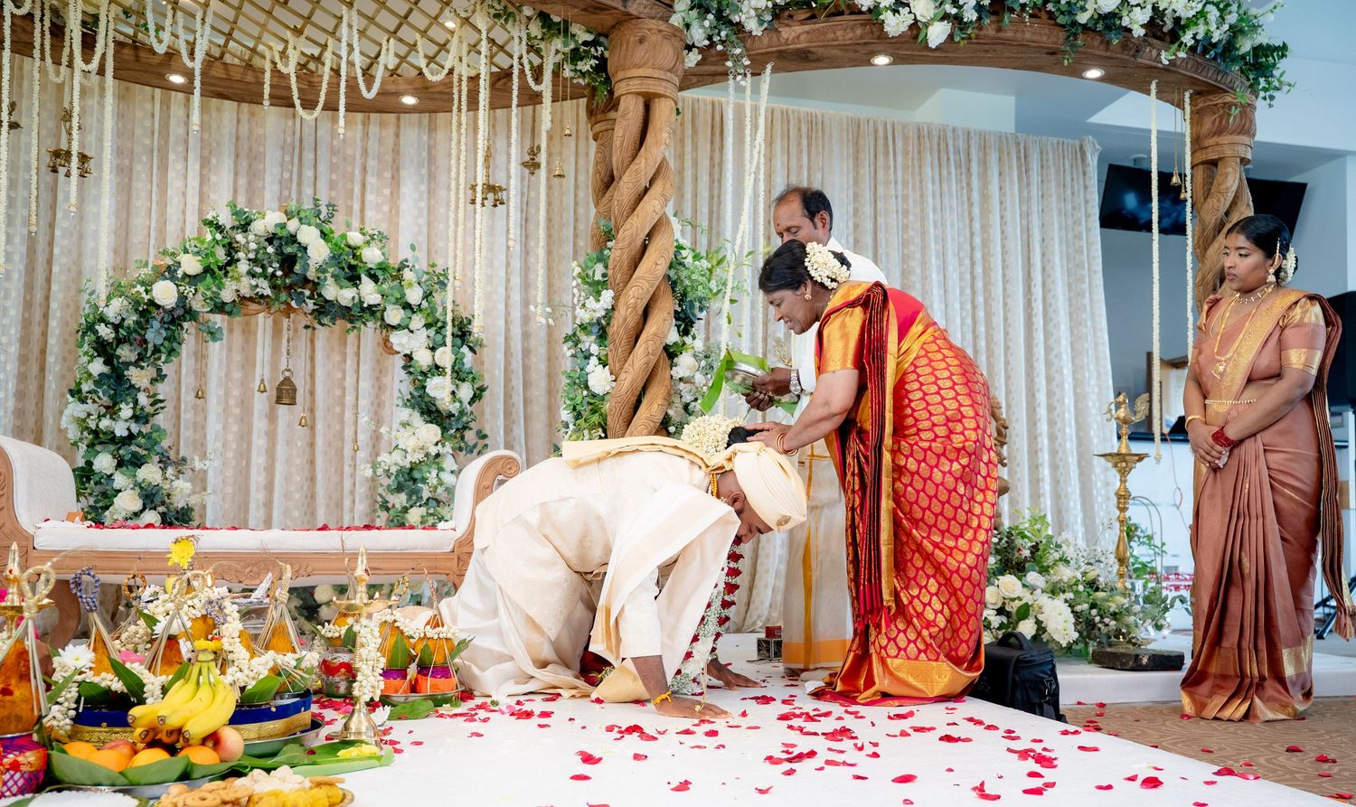 Groom bowing during traditional South Indian Hindu wedding ceremony on flower-petal-strewn white floor with floral arch backdrop.