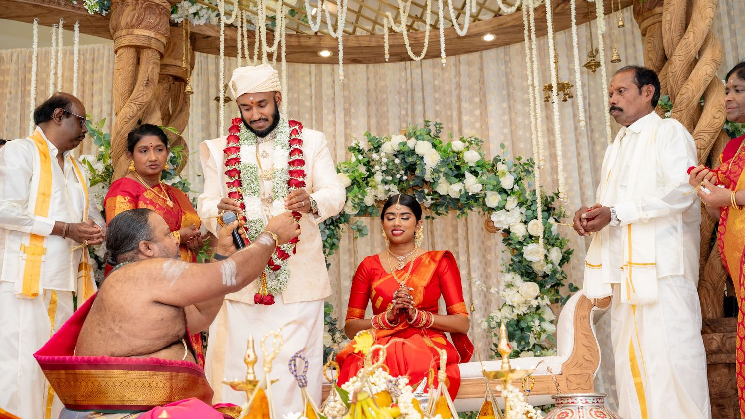 South Indian wedding ceremony with priest blessing couple at ornate mandap decorated with flowers and greenery.