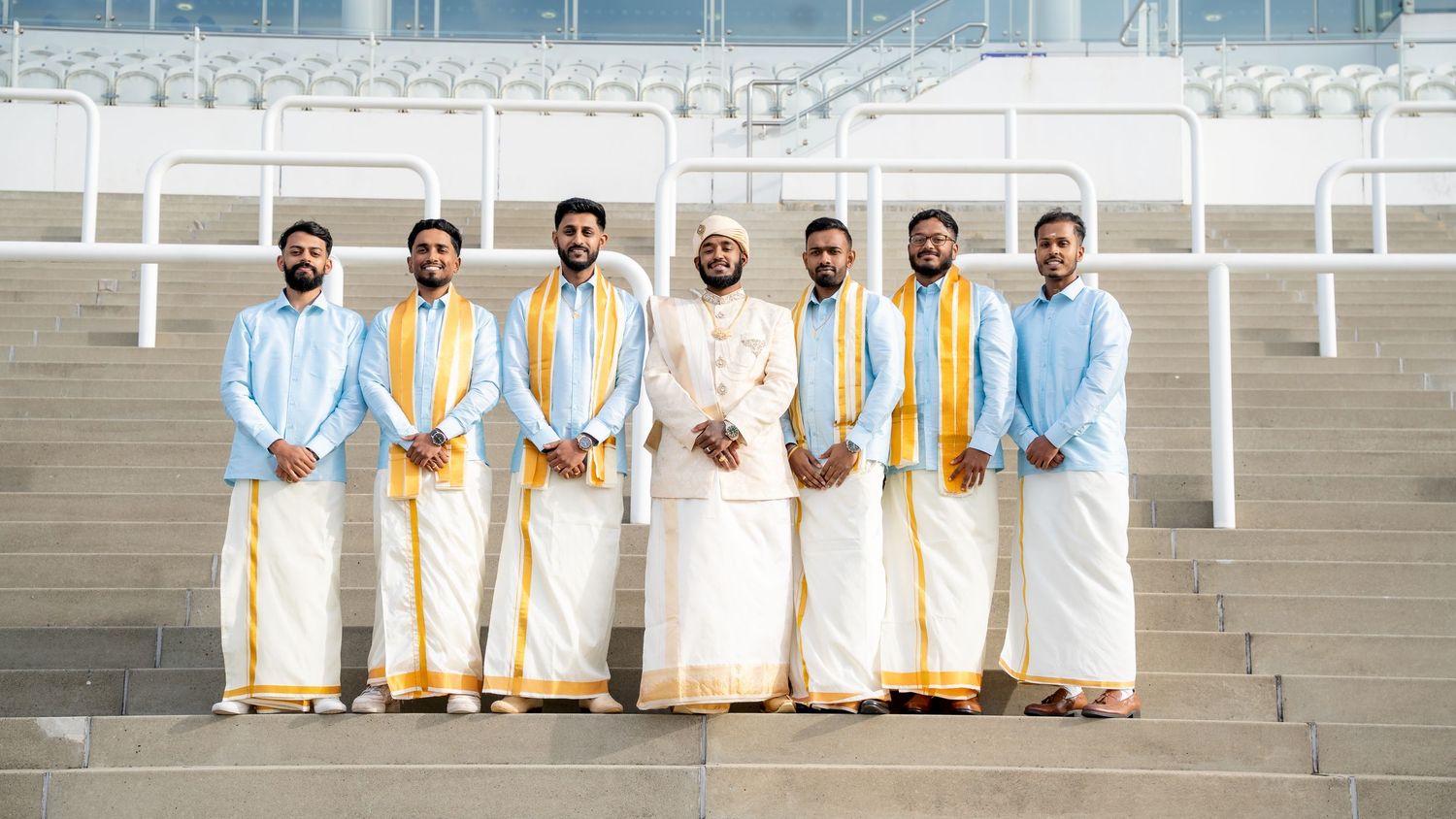 Group of eight men in matching traditional South Asian outfits posing on stadium steps.
