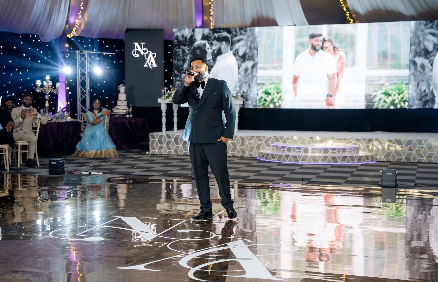 Man in black suit performing at elegant wedding reception with large screen display and decorated venue.