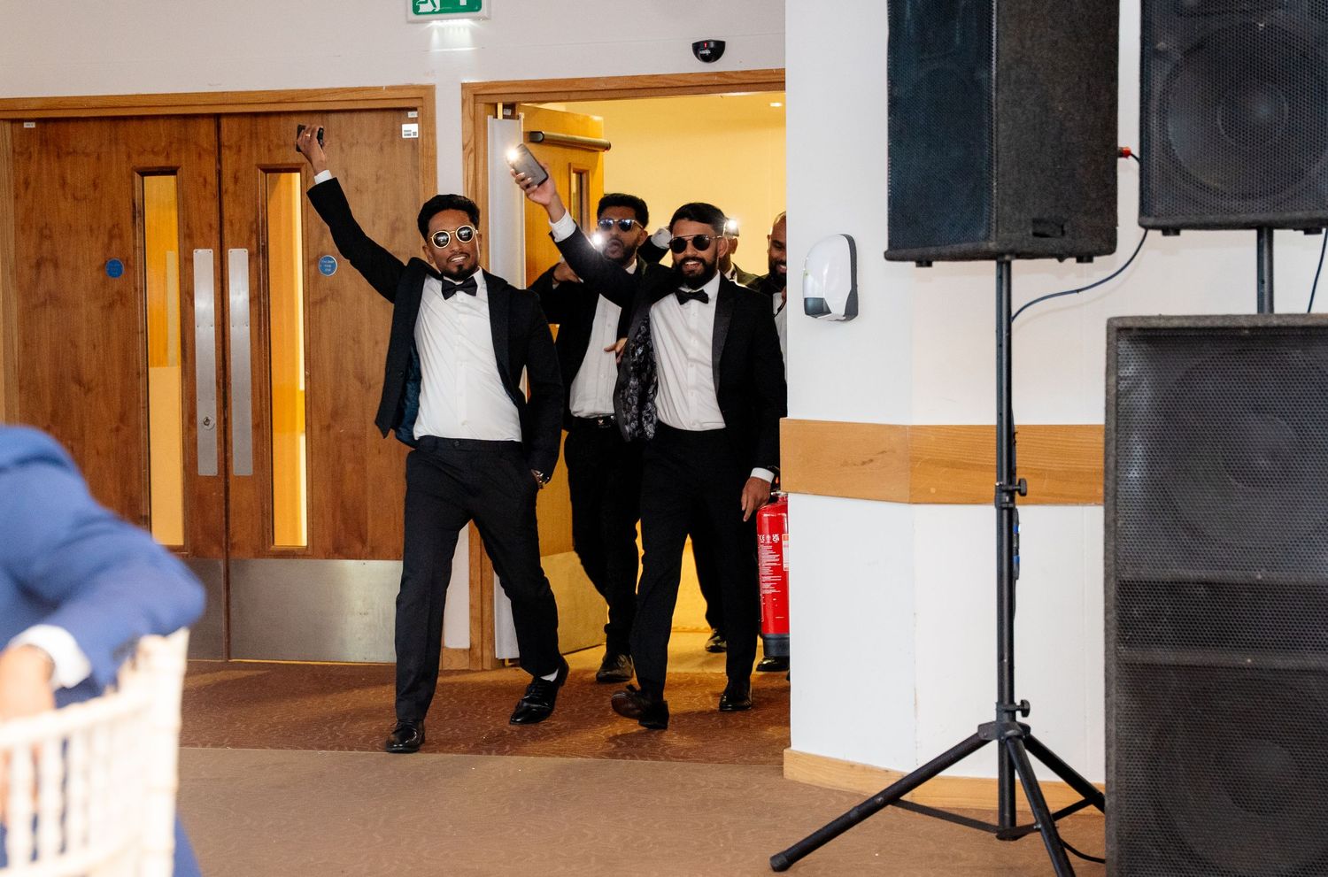 Three men in tuxedos making a celebratory entrance with arms raised at a wedding.