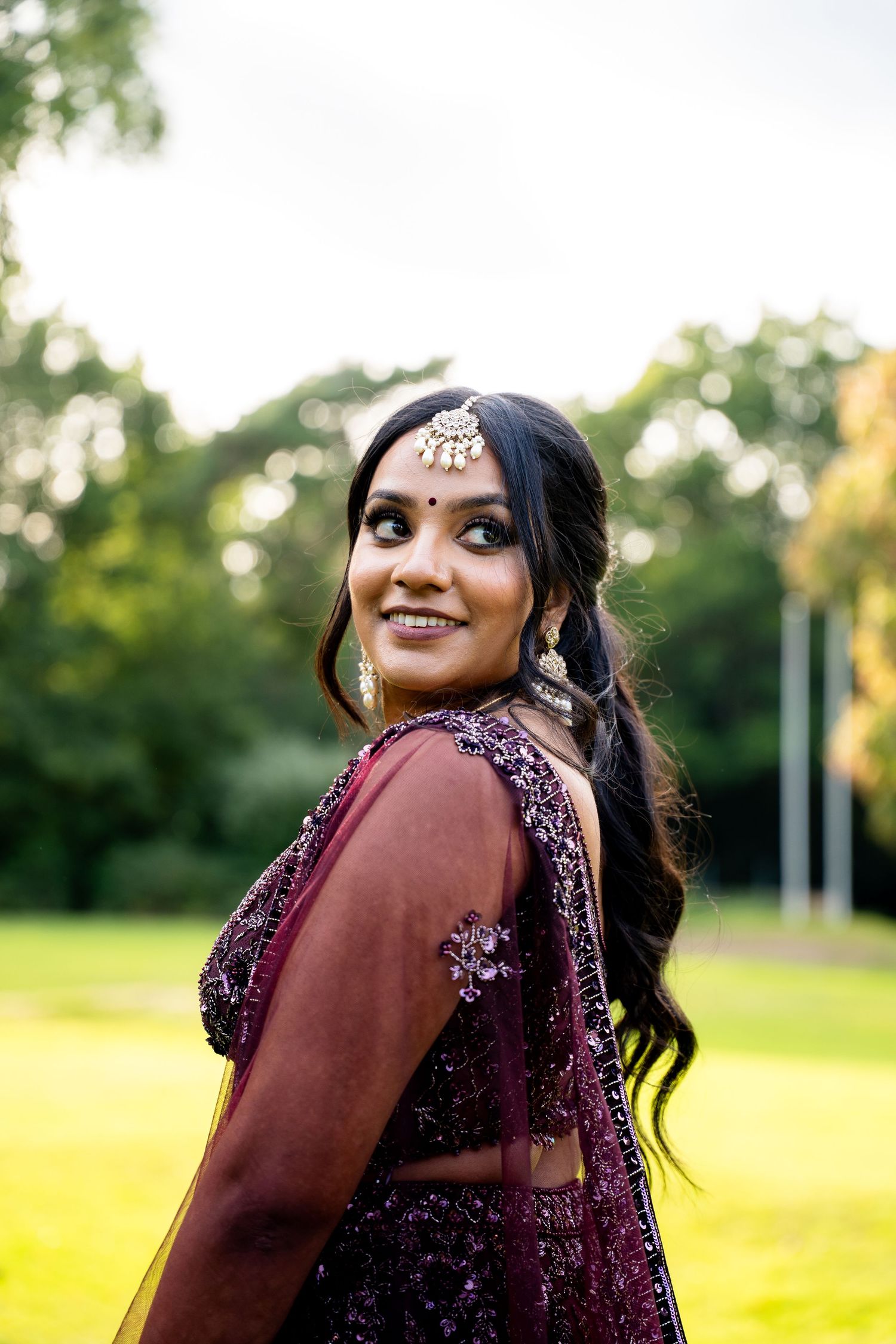 Indian bride in purple embroidered lehenga with maang tikka and chandelier earrings smiling over shoulder in garden setting.