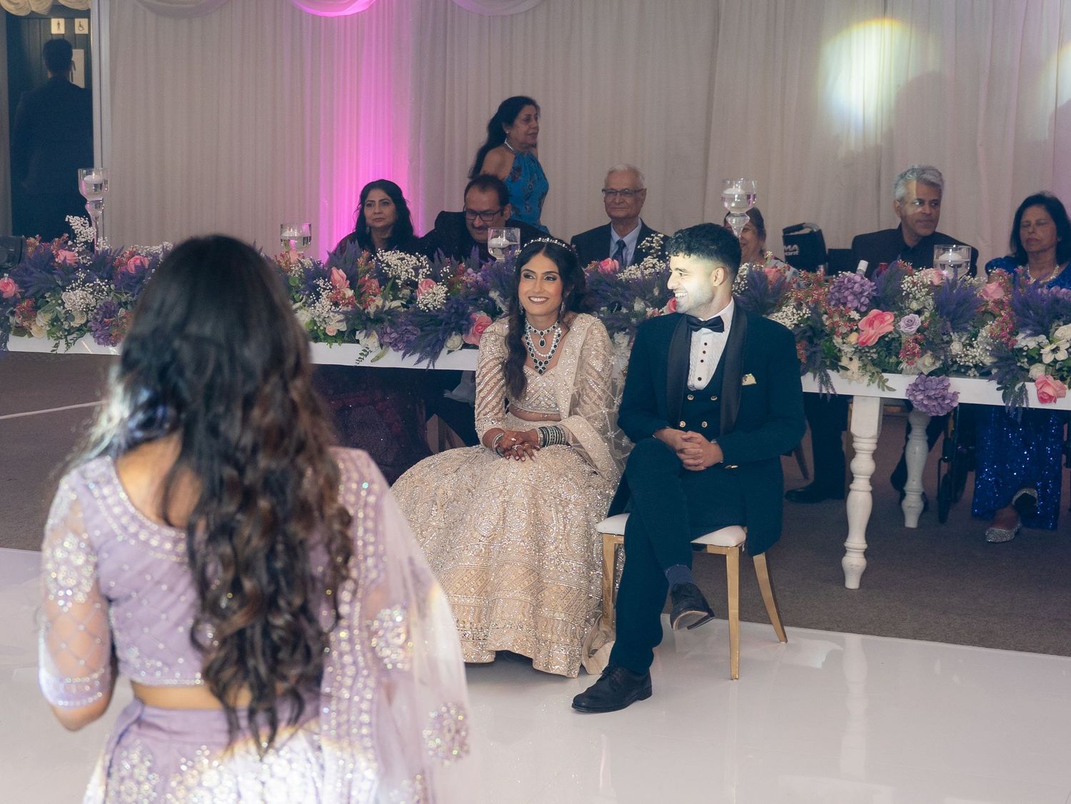 Bride and groom seated at wedding reception head table surrounded by floral arrangements and guests in elegant venue.
