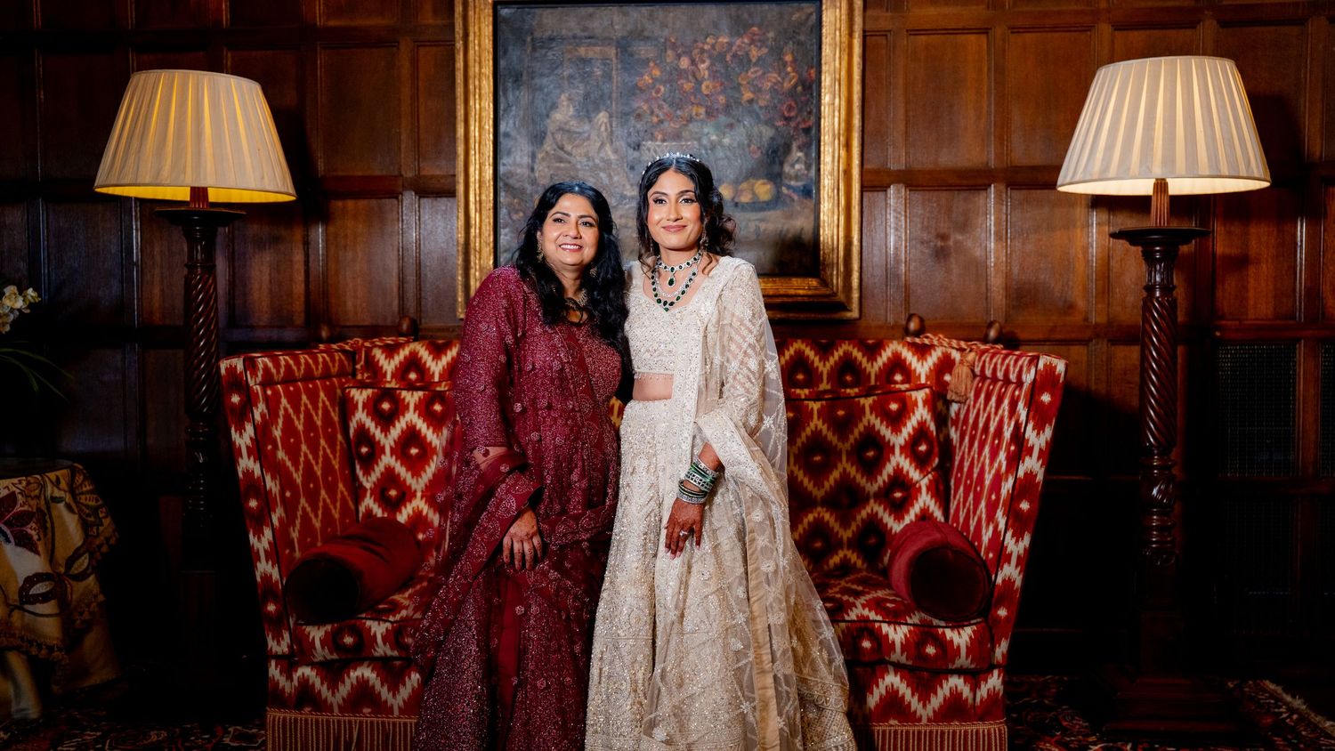 Two women in elegant Indian bridal attire pose together on a red velvet sofa in a luxurious wood-paneled room.