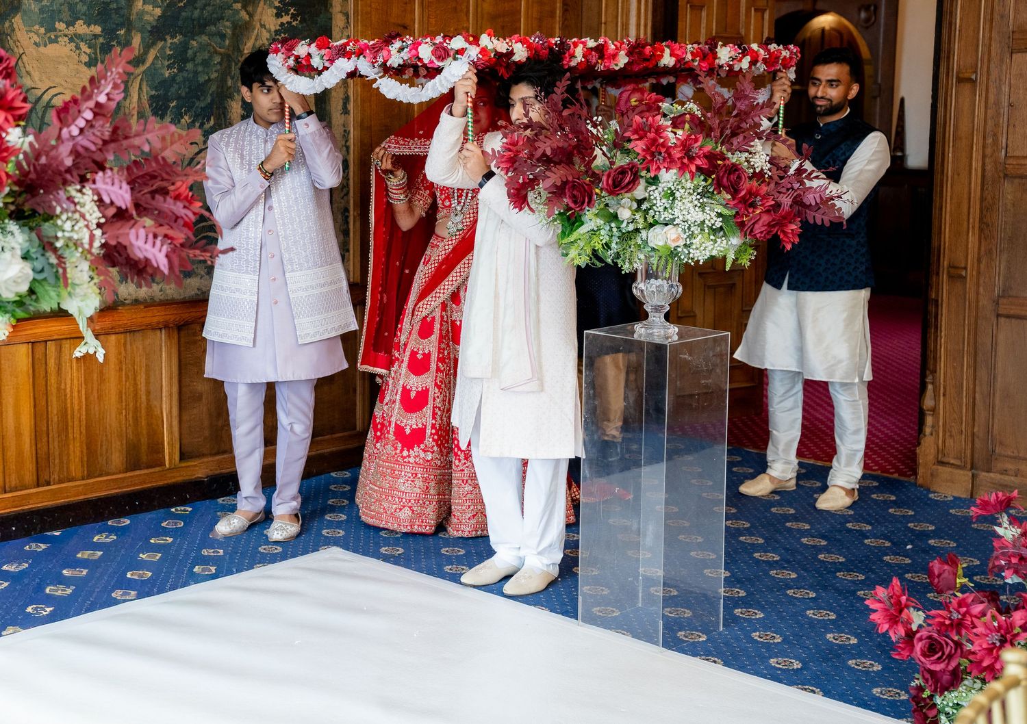 Indian bride in red lehenga walks under floral canopy held by family during wedding ceremony.
