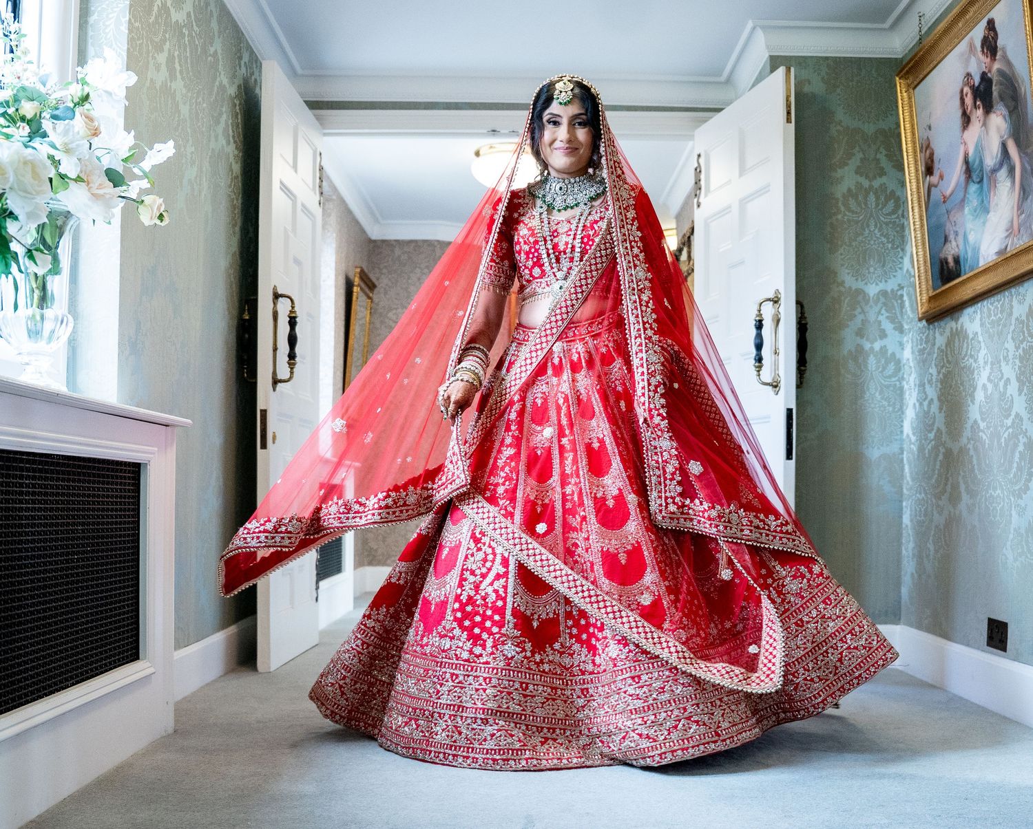 Indian bride twirling in elaborate red embroidered lehenga in elegant hallway setting.