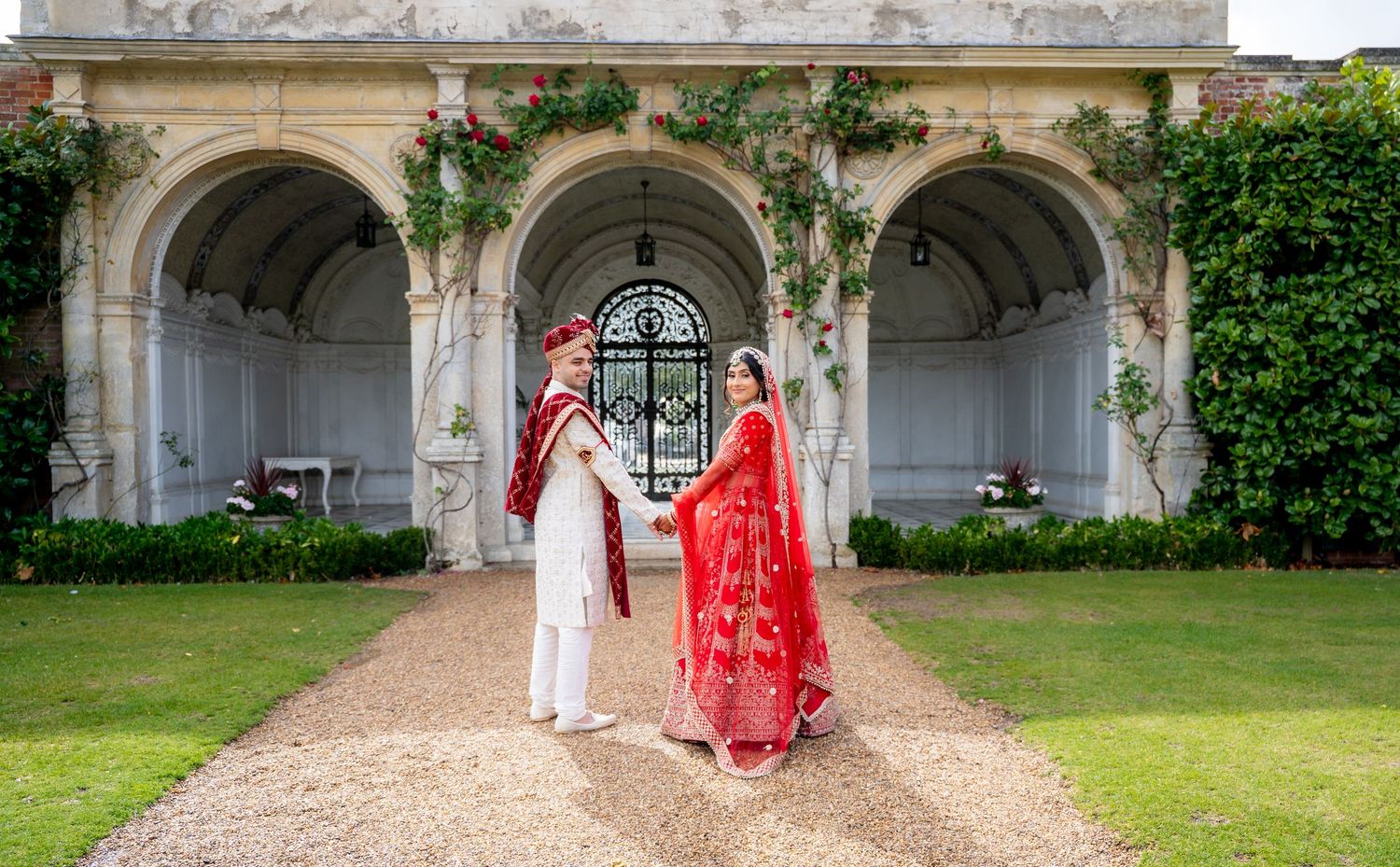 Indian bride and groom in traditional red and white wedding attire posing in an elegant garden with arched stone architecture.