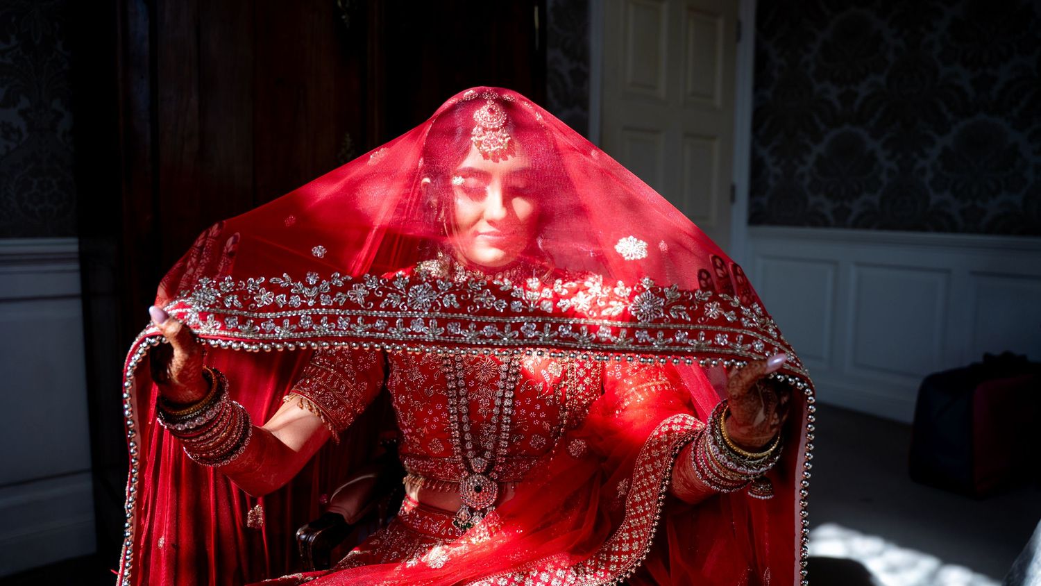 Indian bride in red embroidered lehenga and dupatta veil sitting gracefully in traditional bridal attire.