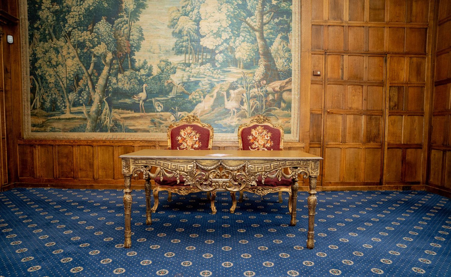Ornate gilded table with red chairs against wood-paneled wall with large tapestry backdrop.
