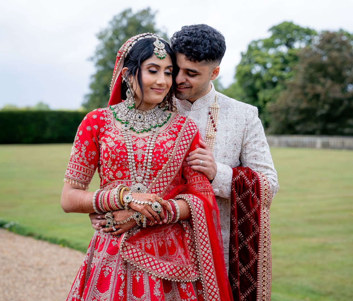 Indian bride in red lehenga and groom in white sherwani embrace outdoors at a garden wedding venue.