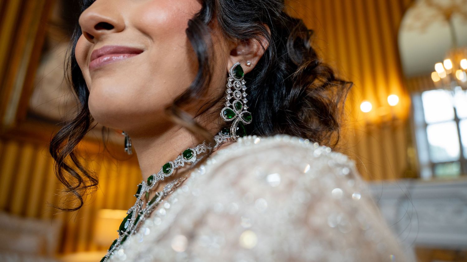 Close-up of Indian bride wearing emerald drop earrings and layered diamond necklace in golden room.