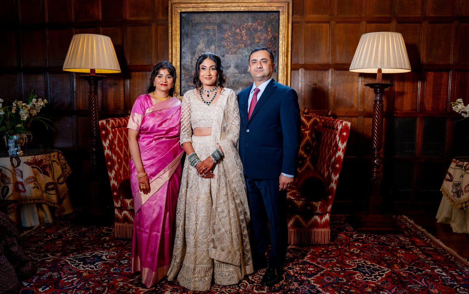 Bride in white lehenga poses with parents in formal Indian attire in a wood-paneled room.