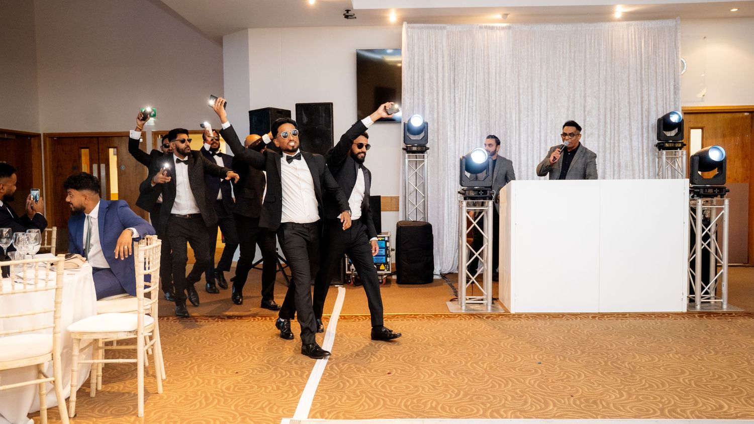 Group of groomsmen in black suits dancing energetically on the dance floor at a wedding reception.