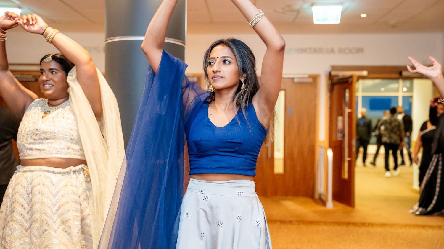 A woman in a blue crop top and skirt dances with arms raised at an Indian wedding celebration.