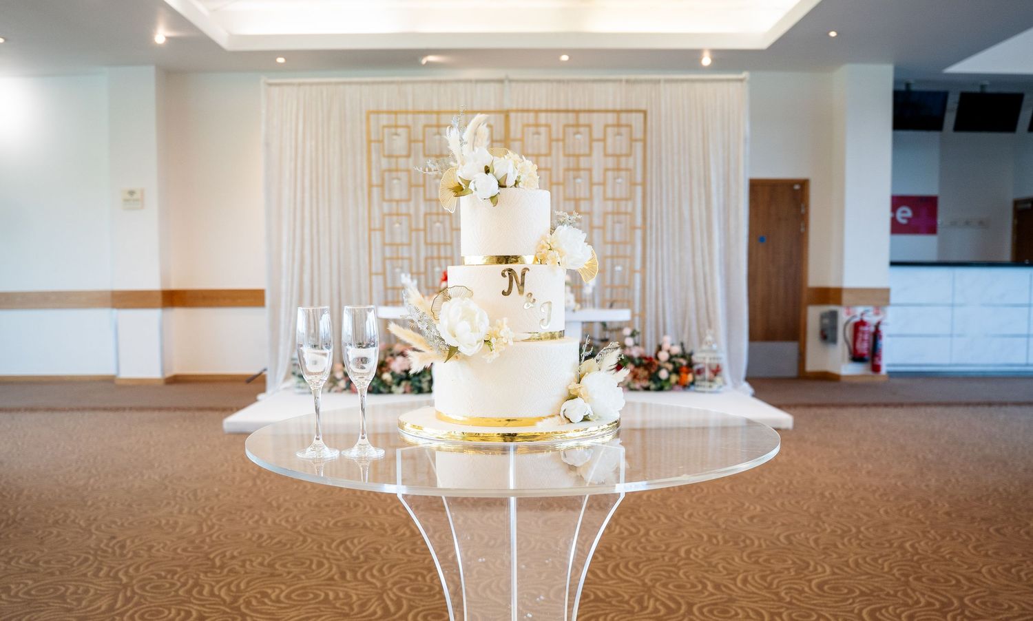 Elegant three-tier white and gold wedding cake displayed on acrylic stand in decorated wedding venue.