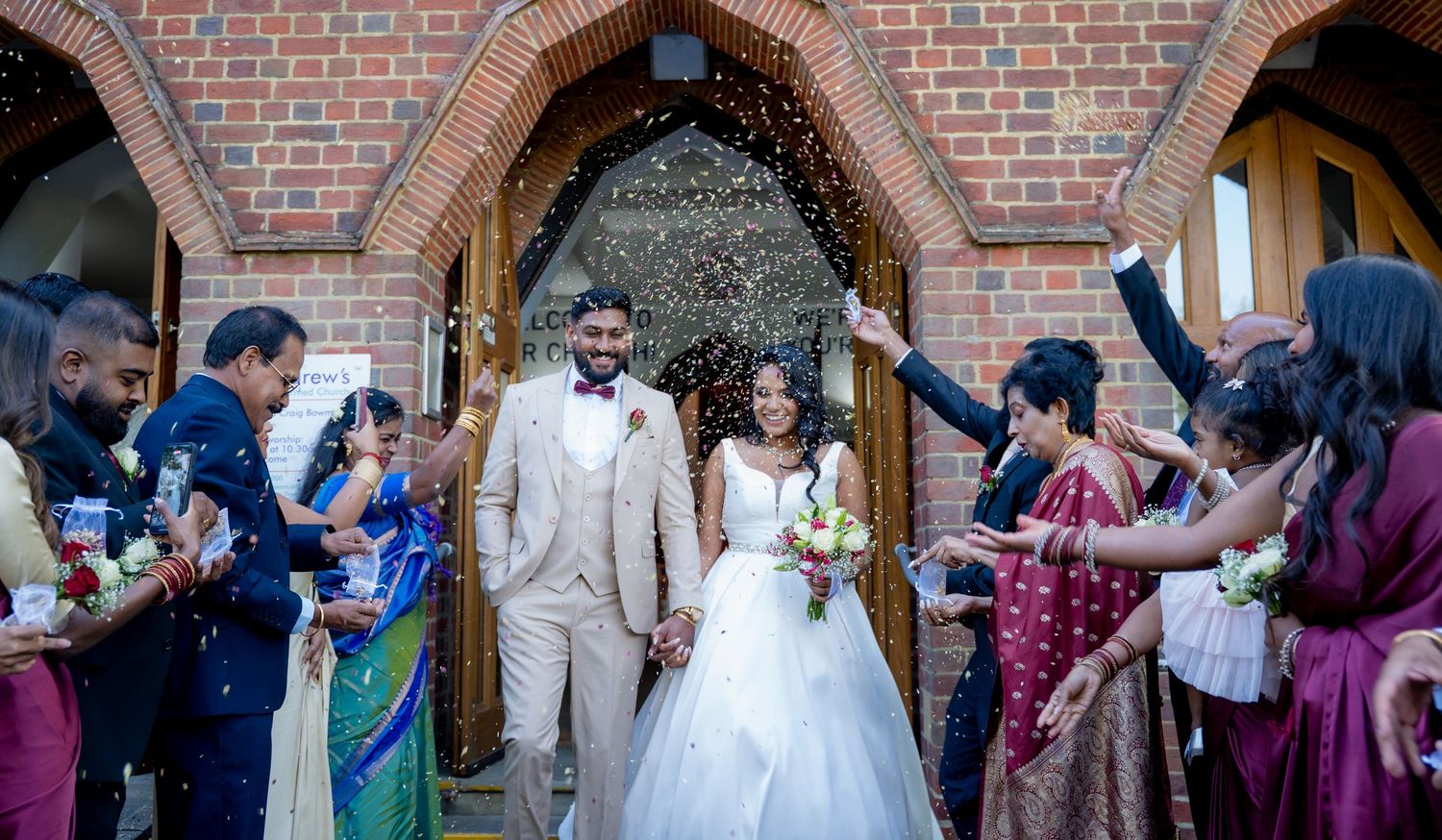 Joyful newlyweds walk through confetti shower outside brick church as guests celebrate their marriage.