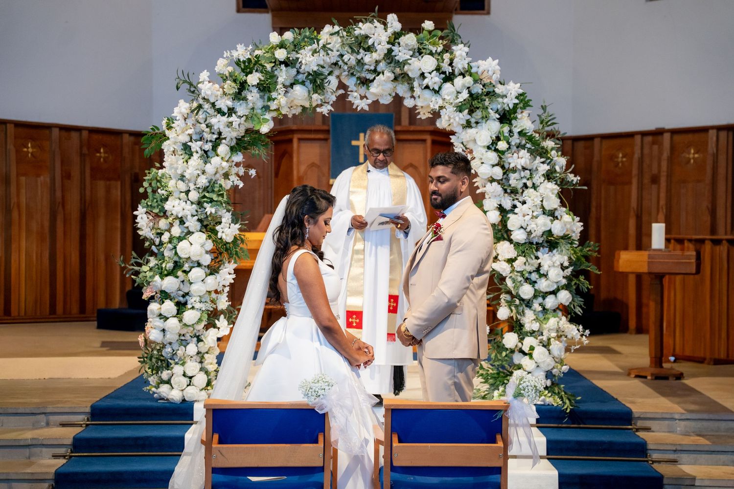 Bride and groom exchange vows under white floral arch with priest at church altar.