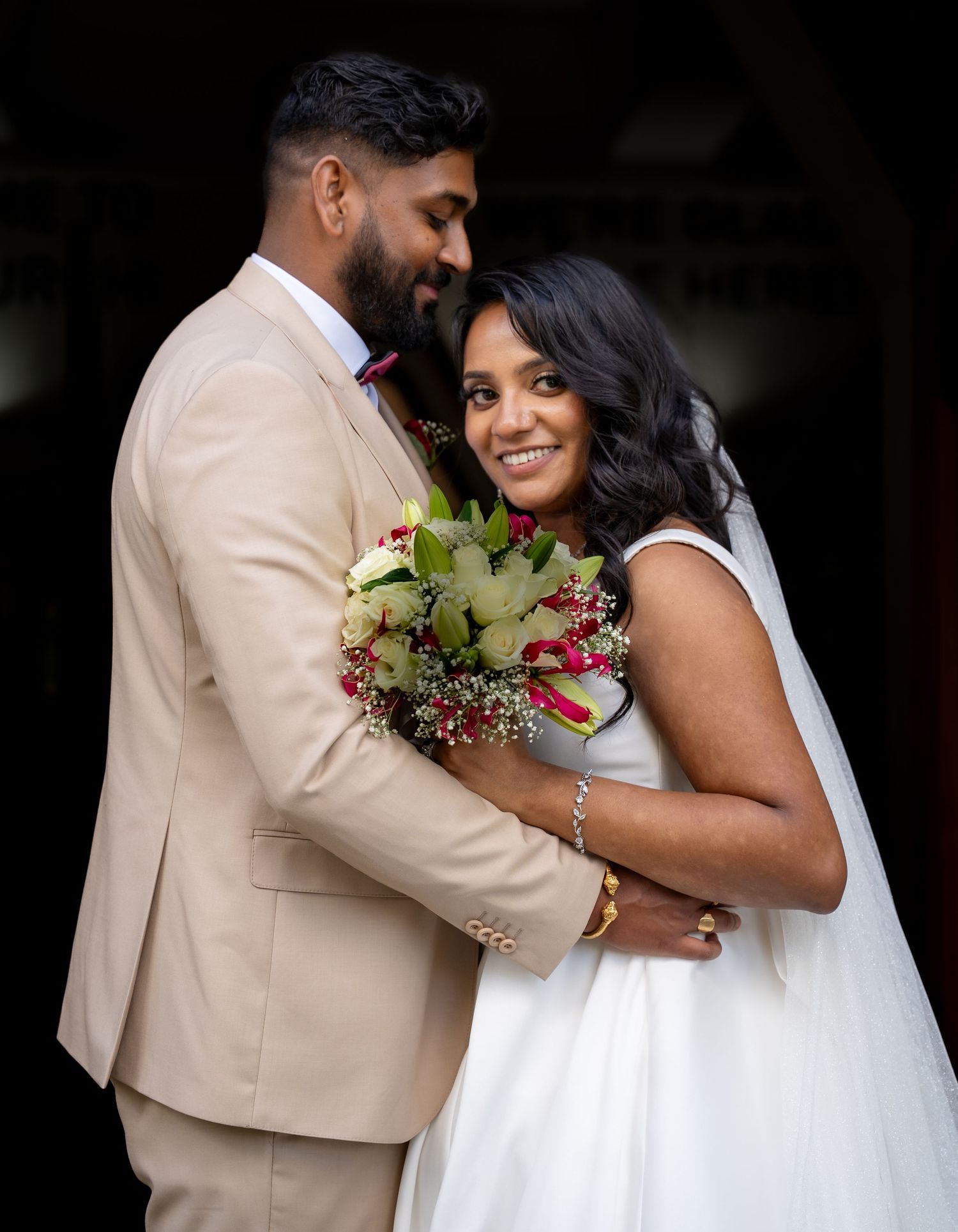 Bride and groom embrace outdoors, she holds red and white bouquet, wearing white dress, he wears beige suit.